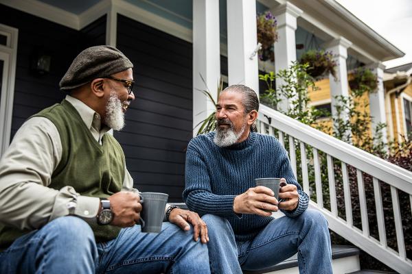 Two men sitting on a porch, holding mugs and smiling at each other